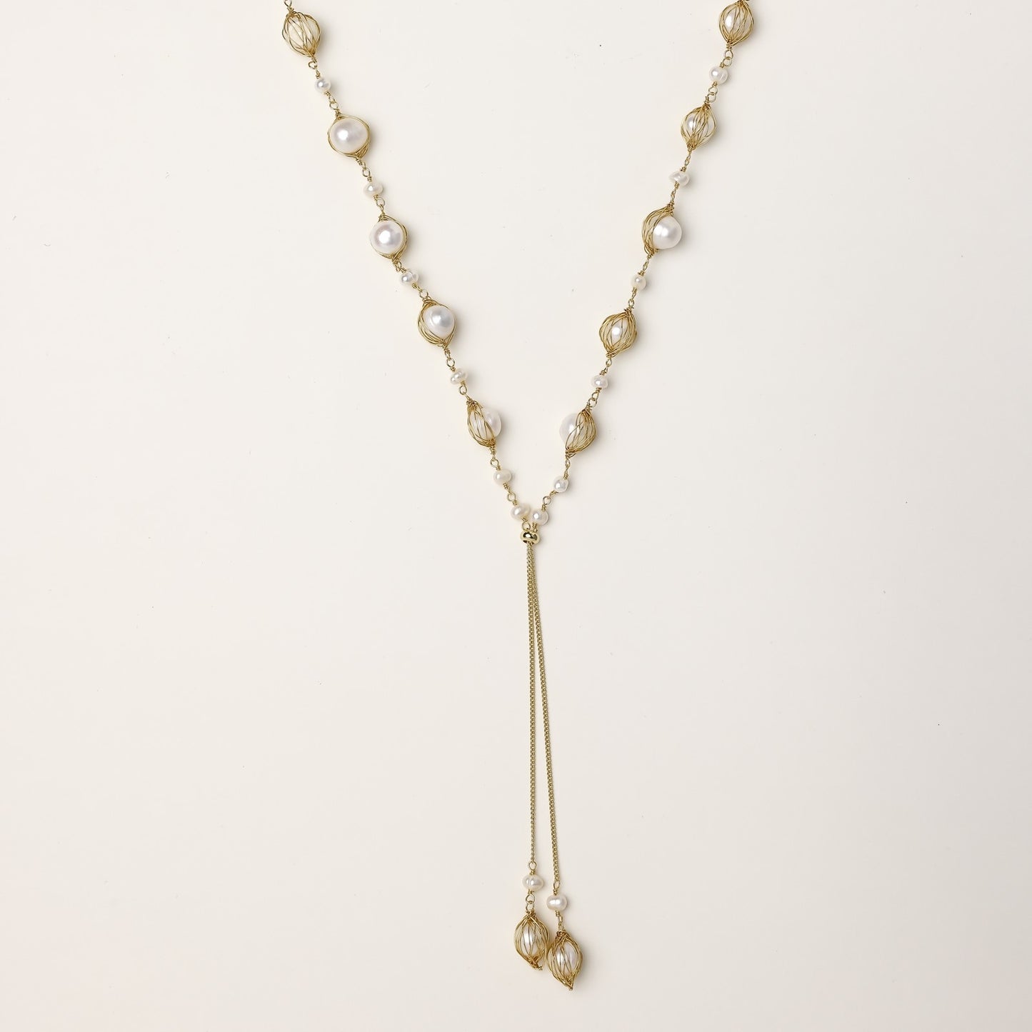 Long gold-plated lariat necklace with wire-wrapped freshwater pearls and chain tassel detail on cream background.