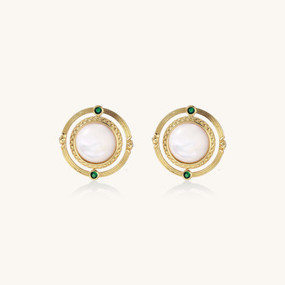 Close-up of gold-plated circular earrings with white mother-of-pearl centers and green zirconia accents on cream background.