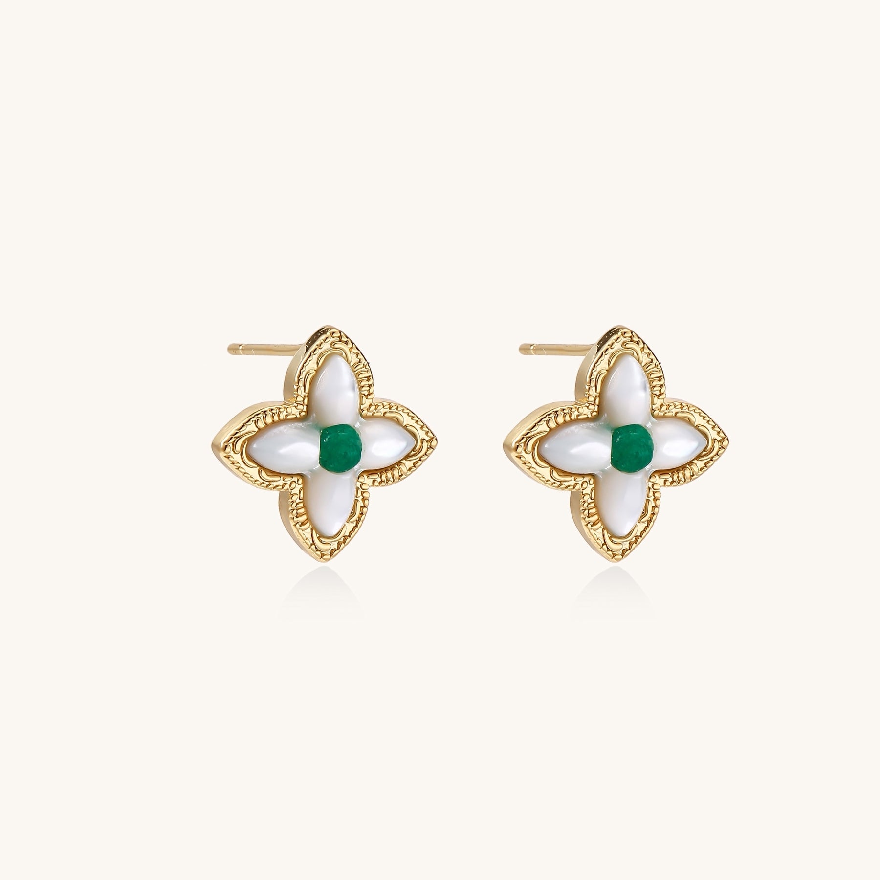 Dainty gold-plated quatrefoil stud earrings with white mother-of-pearl petals and green gemstone centers on beige background.