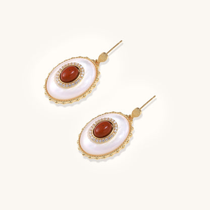 Gold-plated drop earrings with oval mother-of-pearl stones, central red gemstones, and crystal accents on white background.