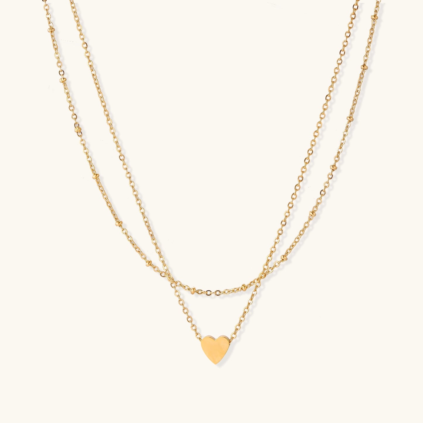 Two-layer gold-plated stainless steel necklace with small heart pendant displayed on beige background