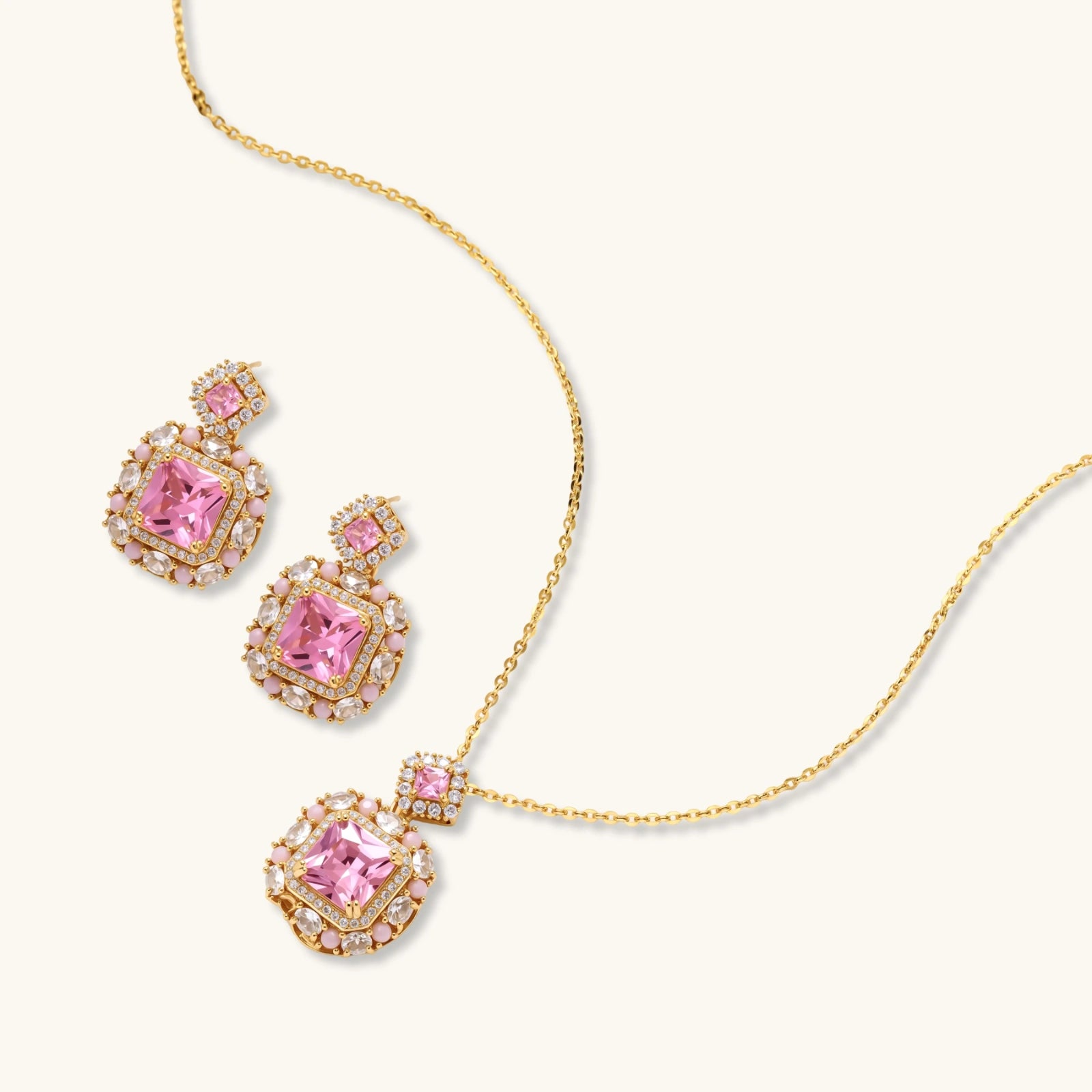 Ornate gold-plated jewelry set with rose pink zirconia center stones and crystal accents on cream background.